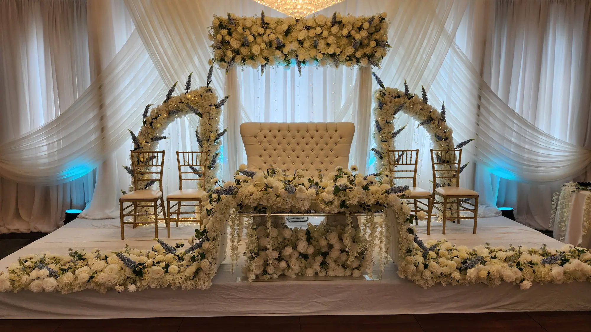 Ivory and gold wedding stage with twin floral arches, a tufted loveseat and a mirrored sweetheart table beneath a crystal chandelier